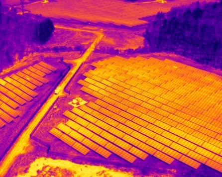 A thermal drone photograph of a solar farm, showing solar panels in various shades of yellow to orange, indicating temperature differences. The surrounding land appears as a dark purple, contrasting with the warmer colors of the panels. This image is used to analyze the temperature of the solar panel surfaces for functionality assessment.