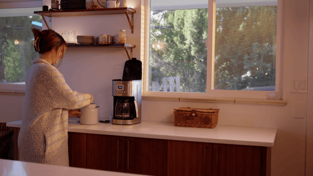 A woman in a furry white robe prepares coffee in her kitchen, standing next to a white countertop. Morning light and kitchen lights create a warm glow.