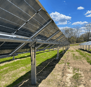 Rows of solar panels mounted on metal frames at a solar farm in Fryeburg, Maine, built using recycled panels, with green grass growing beneath and a bright blue sky with scattered clouds overhead.