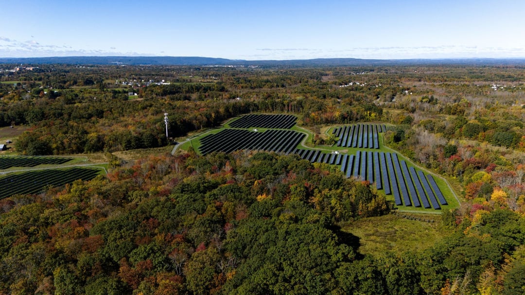Aerial view of a community solar farm in New York with rows of solar panels set among wooded landscape with fall colors and distant hills.