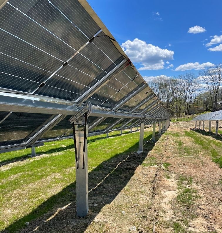 Rows of solar panels mounted on metal frames at a solar farm in Fryeburg, Maine, built using recycled panels, with green grass growing beneath and a bright blue sky with scattered clouds overhead.