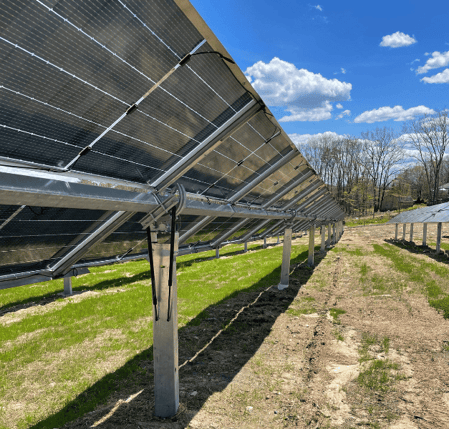 Rows of solar panels mounted on metal frames at a solar farm in Fryeburg, Maine, built using recycled panels, with green grass growing beneath and a bright blue sky with scattered clouds overhead.