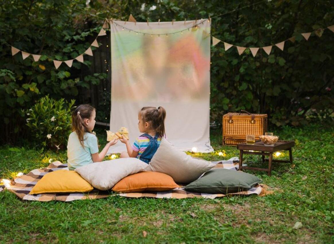 Outdoor movie night setup with two children sitting on cushions and blankets in a grassy yard, eating snacks in front of a white projection screen. Decorative string lights and triangular bunting hang above, with a wicker picnic basket and a small table holding snacks nearby.