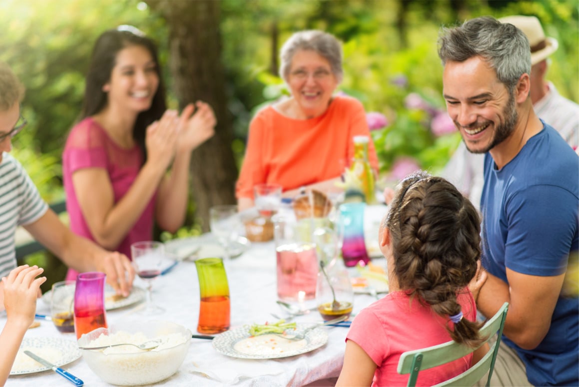 Group of people sitting around an outdoor table enjoying a meal together in a garden setting, with colorful glasses and plates on the table.